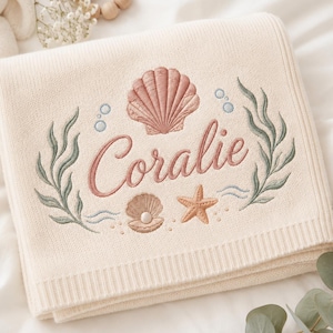 May include: A cream-coloured knit blanket featuring an embroidered design of a large seashell, a smaller pearl-bearing shell, and a starfish. The name Coralie is stitched in pink cursive script, surrounded by green seaweed and blue bubbles.