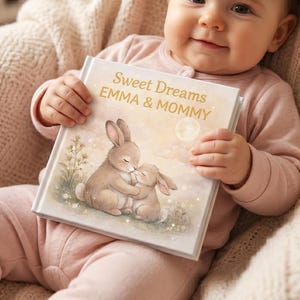 May include: A baby in pink sleepsuit holds a book titled "Sweet Dreams EMMA & MOMMY." The book cover shows a watercolour illustration of two hugging bunnies under a full moon. The baby is seated in a beige armchair.