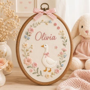 May include: An oval embroidery hoop containing white fabric with a hand-stitched white goose wearing a pink bow, surrounded by a floral wreath of pink, blue, and yellow flowers. The name Olivia is stitched in pink above the goose, and a pink gingham bow is attached to the top.