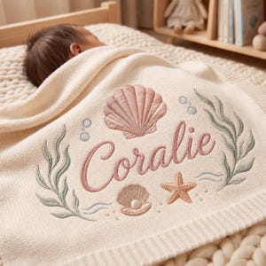 Puede incluir: Una manta de punto para bebé de color crema con un diseño bordado de una concha grande rosa, una concha más pequeña abierta con una perla, una estrella de mar y algas verdes. El nombre Coralie está bordado en cursiva rosa en el centro.