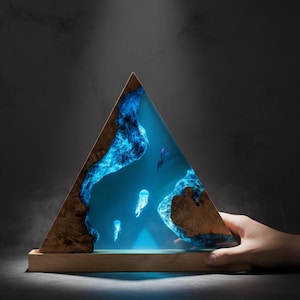 May include: A triangular wooden and resin lamp featuring a marine scene with jellyfish and a diver. The lamp has a wooden base and a glowing blue interior, creating an underwater effect. The dimensions are approximately 10 inches tall and 8 inches wide.