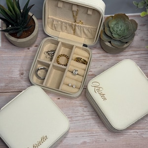 May include: Open white jewelry travel case with multiple compartments, holding rings, earrings, and necklaces. Two closed cases with personalized names are also visible. The cases are on a wooden surface, with small potted plants in the background.