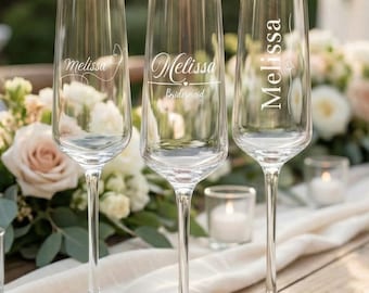 Peut inclure: Trois flûtes à champagne transparentes gravées du nom "Melissa". L'une des flûtes porte également le mot "Bridesmaid". Les flûtes sont posées sur une table en bois avec un tissu blanc et des fleurs en arrière-plan.