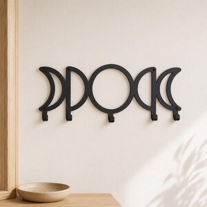 May include: A matte black metal wall-mounted hook rack featuring a horizontal design of the lunar cycle phases. The piece displays five distinct moon shapes, including crescents and a central full circle, each equipped with a small downward hook.