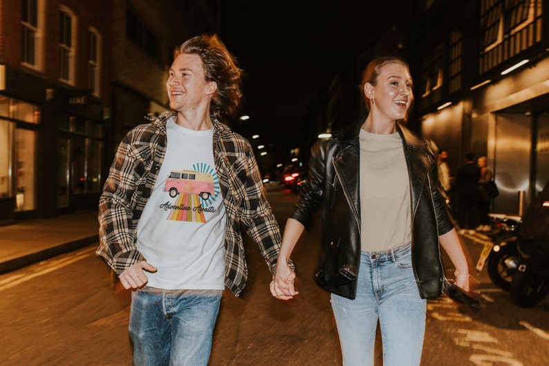 May include: A couple walking down a city street at night. The man is wearing a white t-shirt with a colorful graphic of a van and the text "Adventure Awaits". The woman is wearing a beige t-shirt under a black leather jacket.