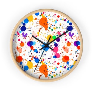 May include: A round wall clock with a white face and a wooden frame. The clock face features a colorful paint splatter pattern in shades of blue, green, yellow, orange, pink, and purple.