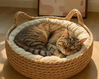 Pode incluir: Um gato malhado dormindo em uma cama redonda para gatos feita de material tecido. A cama tem uma almofada creme e alças em forma de orelha. O gato é marrom com listras pretas.