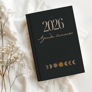 May include: Black 2026 lunar agenda notebook with gold lettering and moon phase illustrations. The cover features the year 2026 and the words "Agenda lunaire". The notebook is lying on a white surface with dried flowers.