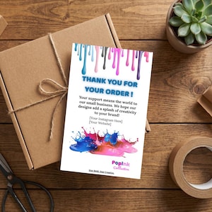 Op de afbeelding: Een witte bedankkaart met blauwe, roze en zwarte verfdruppels bovenaan. De kaart luidt "DANK U VOOR UW BESTELLING!" en bevat een kleurrijk verfspatontwerp. Een bruine kartonnen doos en een vetplant staan op de achtergrond.
