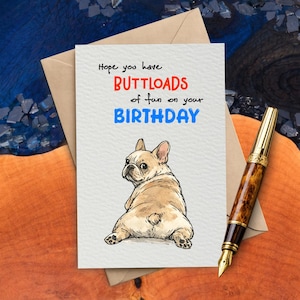 May include: Birthday card with a French Bulldog illustration. The card reads "Hope you have BUTTLOADS of fun on your BIRTHDAY" in red and blue text. A gold and brown pen rests on the wooden surface.