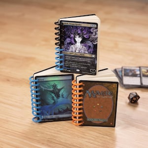 May include: Three small notebooks constructed from Magic: The Gathering trading cards bound with colorful plastic spiral coils. The top notebook features Ashiok, Dream Render, while the bottom two show an Island card and the game's card back.