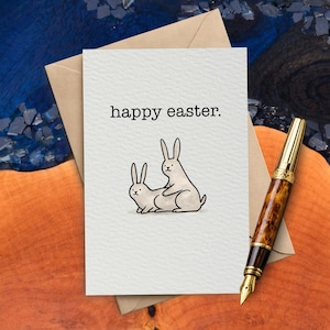 May include: A greeting card with the text "happy easter." features a humorous illustration of two rabbits. The card is on a beige envelope, with a gold and brown pen resting on a wooden surface.