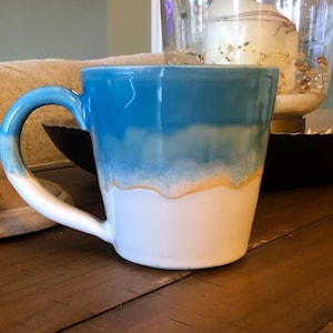 May include: A ceramic mug with a blue and white gradient design, accented with a gold band. The mug has a curved handle and a glossy finish. The mug is a kitchen and dining item.