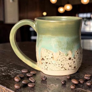 May include: A ceramic mug with a curved handle featuring a two-tone glaze. The upper portion is a smooth sage green that transitions into a speckled cream base with visible drip patterns. The mug is surrounded by scattered roasted coffee beans.