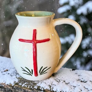 May include: A cream-colored ceramic mug with a rounded body and a single handle. The front features a raised, hand-painted red cross design positioned above small, dark green leaf-like markings. The interior of the mug has a light green glaze.