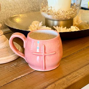 May include: A round, light pink ceramic mug with a glossy finish and a smooth, curved handle. The exterior of the mug features a raised, embossed cross design centered on the front. The interior of the mug is a light, neutral beige color.