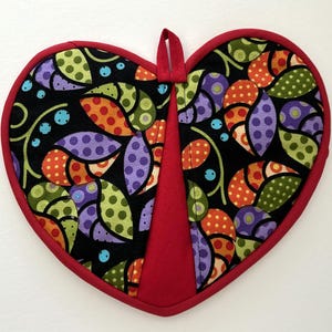 May include: Heart-shaped quilted potholder with a red border and hanging loop. The black fabric is patterned with colorful leaves in orange, green, and purple with polka dots. The center is solid red.