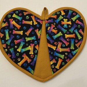 May include: Heart-shaped pot holder with a black background featuring colorful rainbow-colored dog bones and paw prints. The pot holder has a gold-colored border and a loop for hanging.