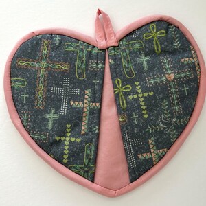 May include: Heart-shaped quilted potholder with a pink border and hanging loop. The dark gray fabric features a pattern of crosses in various styles, with green and white accents. A pink fabric panel divides the potholder.