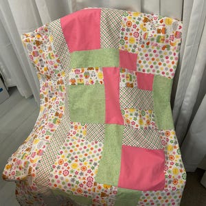 Puede incluir: Un edredón de retazos con una mezcla de patrones y colores. El edredón presenta grandes bloques rosas y verdes, rodeados de tela con estampados florales, de lunares y de animales. El edredón está sobre una silla.