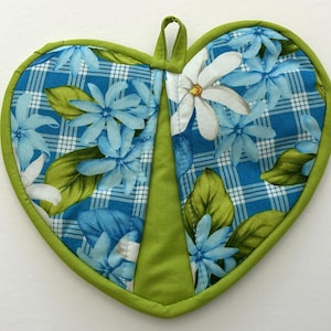 May include: Heart-shaped quilted potholder with a floral pattern. The design features blue flowers and green leaves on a blue and white plaid background. The potholder has a green border and a loop for hanging.