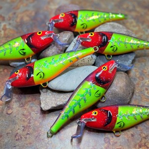 May include: Six colorful fishing lures with red heads, chartreuse bodies, and silver accents are arranged on a bed of rocks. Each lure has a clear plastic lip and a small hook. The lures are designed for catching fish.