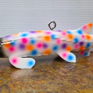 May include: A colorful, decorative fish-shaped ornament. The white body is speckled with pink, purple, orange, and blue dots. The head is black with a green eye, and the fins are tipped with orange and blue. A silver loop is attached for hanging.