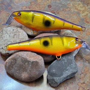 May include: Two yellow and orange fishing lures with black spots and clear plastic fins. The lures are resting on a bed of smooth, rounded stones. The lures have gold-colored hooks.