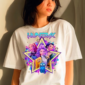 May include: White t-shirt with a graphic of three animated women inside a star, with the word "HUNTRAX" above them. The graphic has purple, blue, and yellow accents. A blue cartoon creature is also featured in the graphic.