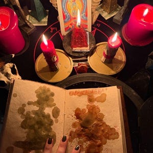 May include: A mystical scene featuring lit red candles, a tarot card, and an open book with the word "CABRACADABRA" visible. The scene is set on a black surface with other ritualistic objects.