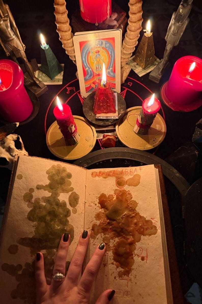 May include: A mystical scene featuring lit red candles, a tarot card, and an open book with the word "CABRACADABRA" visible. The scene is set on a black surface with other ritualistic objects.