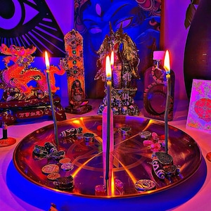 May include: A round metallic tray featuring an engraved pentagram, holding three lit thin green candles and a central clear crystal point. Scattered around the tray are various small stone beads, metallic coins, and patterned cylindrical charms.