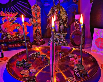 Puede incluir: Una bandeja metálica redonda con un pentagrama grabado, que sostiene tres velas verdes delgadas encendidas y una punta de cristal transparente central. Alrededor hay varias cuentas de piedra, monedas metálicas y amuletos cilíndricos.