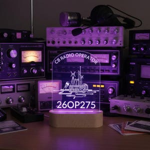 May include: An arched acrylic sign with a light-up base, featuring the text "CB RADIO OPERATOR" and "26OP275" alongside a graphic illustration of a radio, microphone, and antennas. The sign emits a bright purple glow against a background of equipment.