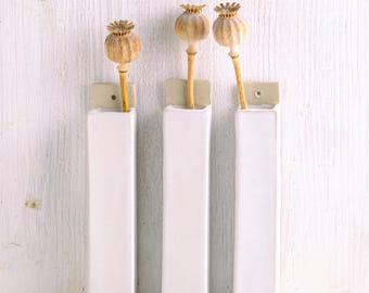 May include: Three white ceramic wall-mounted bud vases, each rectangular in shape with a flat back and a small mounting tab at the top. Each vase holds a single dried poppy seed pod on a thin, light brown stem, creating a vertical arrangement.