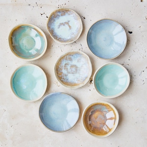 May include: An overhead shot of eight small, handmade ceramic bowls in various shades of blue, turquoise, and brown. The bowls have a rustic, textured appearance with glazed interiors and rims. The arrangement is circular, showcasing the different colors and textures.