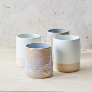 May include: Four ceramic tumblers in different colours. Two are white with beige bases, one is light brown with blue and purple hues, and the last is light blue. The tumblers are cylindrical with a smooth finish.