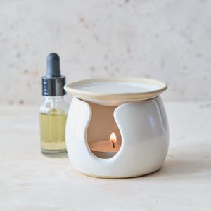 Op de afbeelding: Een witte keramische oliebrander met een ronde vorm en een brede schaal aan de bovenkant, met een brandend waxinelichtje in de opening. Naast de brander staat een klein doorzichtig glazen flesje met een zwarte druppeldop en gele vloeistof.