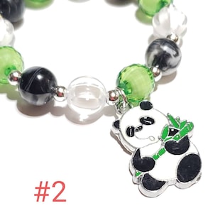 May include: A green, black, and white beaded bracelet with a panda bear charm holding a piece of bamboo. The charm is silver with black and white enamel. The bracelet has a clear bead and the text #2.