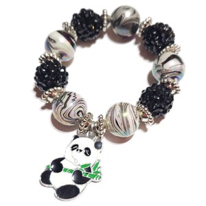 May include: A black and white panda charm hangs from a bracelet made of black and white beads. The beads are a mix of small black beads and larger white beads with black swirls.