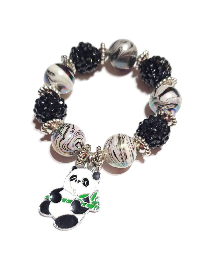 May include: A black and white panda charm hangs from a bracelet made of black and white beads. The beads are a mix of small black beads and larger white beads with black swirls.