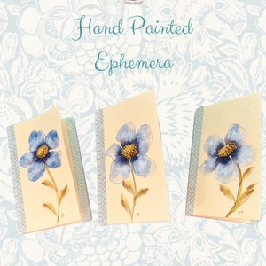 May include: Three hand-painted cards with blue flowers on a cream background. The cards are trimmed with a gold and blue patterned border.