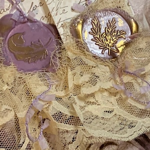 May include: Two vintage-style tags with purple wax seals and gold accents. The seals feature a feather and a sprig of leaves. The tags are tied together with a delicate lace ribbon and a gold safety pin.
