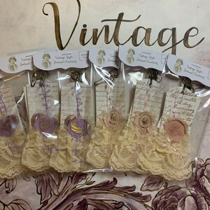May include: Five vintage style journal embellishments with lace, wax seals, and a handwritten note. The embellishments are packaged in clear plastic bags and feature a purple and gold color scheme.