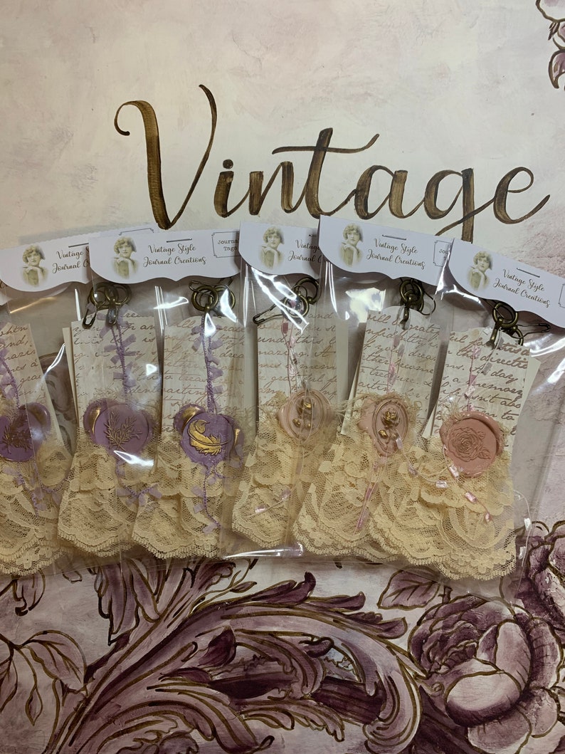 May include: Five vintage style journal embellishments with lace, wax seals, and a handwritten note. The embellishments are packaged in clear plastic bags and feature a purple and gold color scheme.