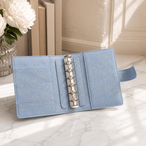 May include: A light blue six-ring binder with a textured finish, featuring a silver metal ring mechanism in the center, multiple interior card slots on the left side, and a small side tab closure with a snap button fastener.