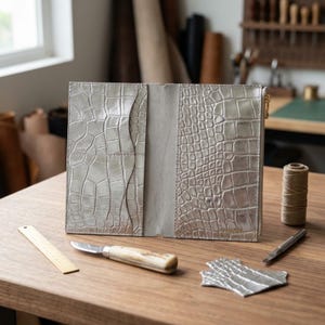 May include: A metallic silver leather wallet featuring a textured crocodile skin pattern. The interior displays multiple card slots on the left side and a zippered compartment on the right. The wallet is open, revealing a smooth, light gray suede lining.