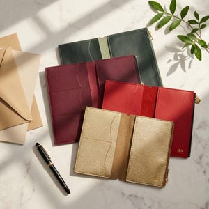 Puede incluir: Cuatro fundas para pasaporte abiertas en verde oscuro, burdeos, rojo y dorado metalizado. Cada funda cuenta con ranuras para tarjetas y un bolsillo lateral con cremallera. La funda roja tiene grabadas las letras IRN y la dorada también muestra IRN.