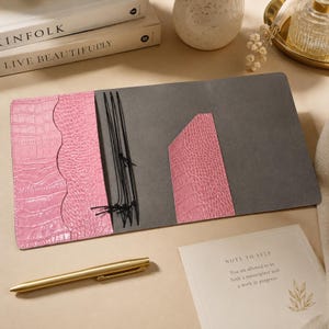 Könnte beinhalten: Ein Notizbuchumschlag aus rosa Leder mit Krokodilprägung, grauem Innenfutter, mehreren Fächern und schwarzen Gummibändern zur Befestigung von Einlagen. Der Umschlag liegt flach neben einem goldfarbenen Metallstift und einer Karte mit Text.