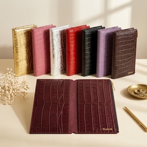 Puede incluir: Una colección de carteras rectangulares con textura de cocodrilo, disponibles en oro, rosa, plata, rojo, negro, lavanda y marrón oscuro. Una cartera marrón oscuro está abierta, mostrando ranuras para tarjetas y el nombre Mahtub impreso.